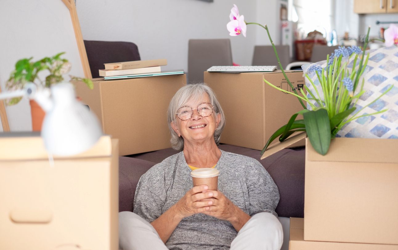 Older woman seated on the floor with a coffee cup surrounded by moving boxes.