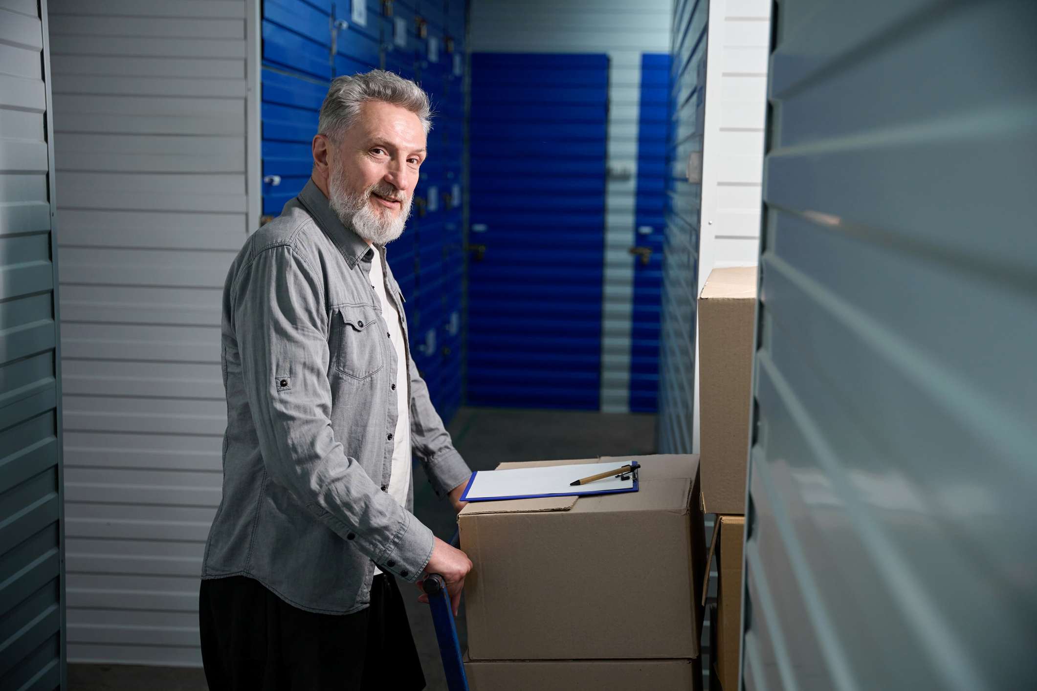 Adult male standing near big trolley with stacked boxes in storehouse and looking at camera