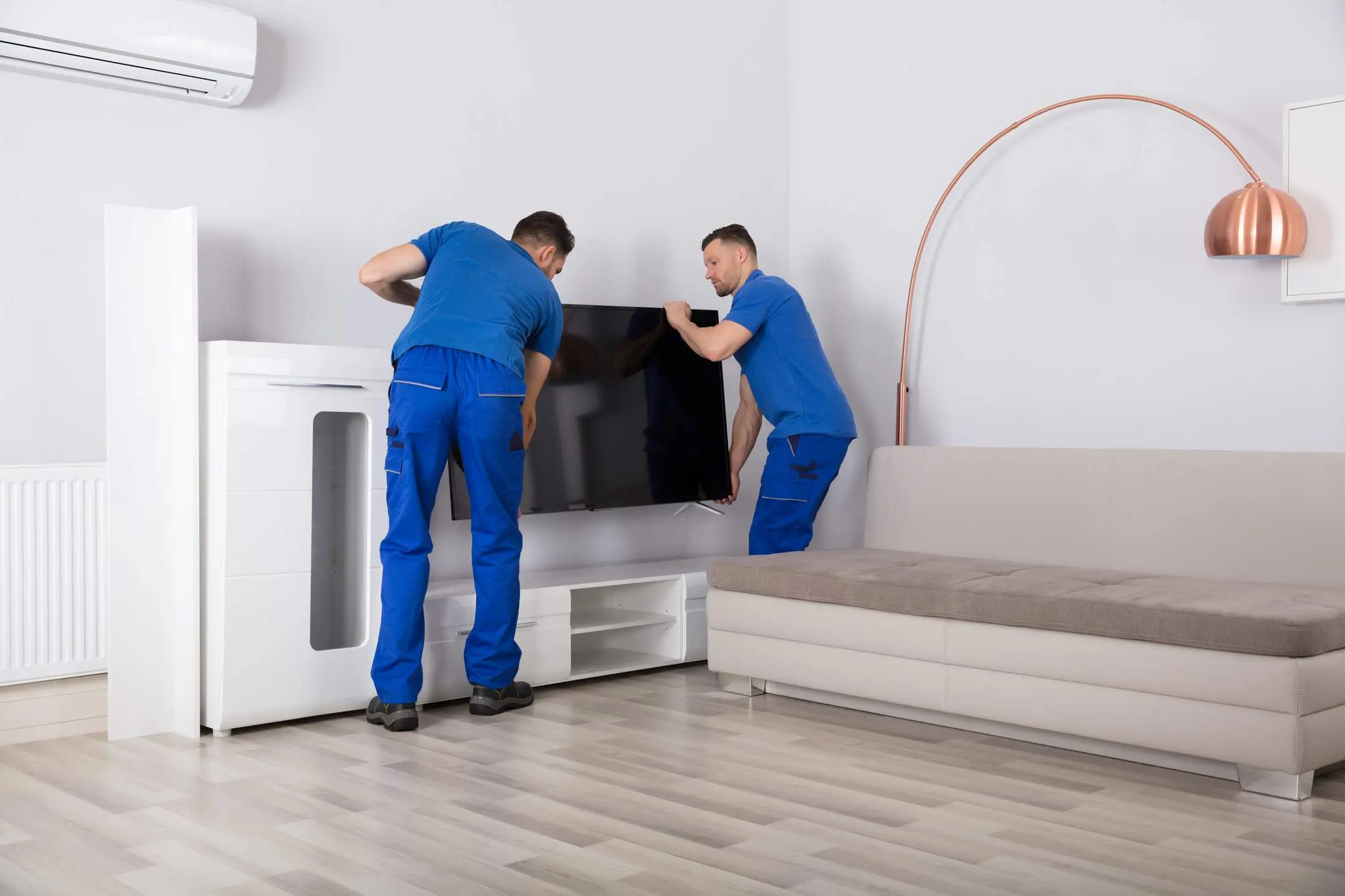 Two Young Male Movers In Uniform Placing Television In Living Room