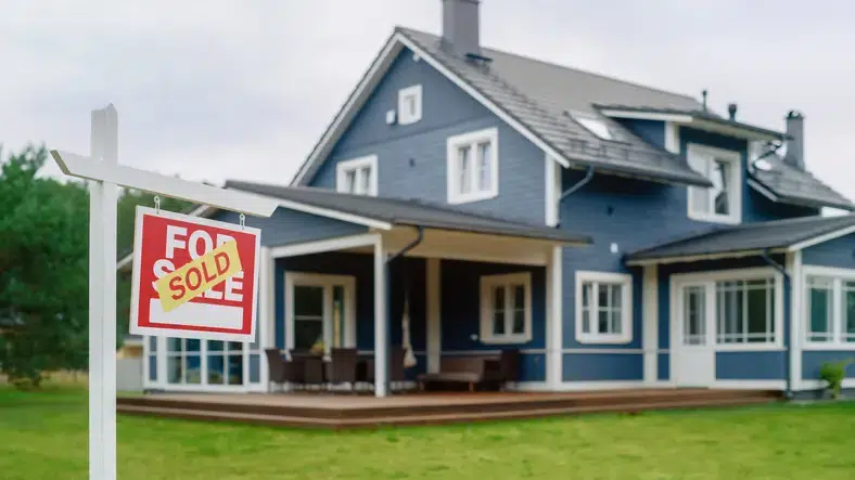 Red Property Sold Sign in a Garden in Front of a Big Blue Stylish House with Traditional Architecture. Hot Housing Market Concept with Residential Home in the Suburbs.