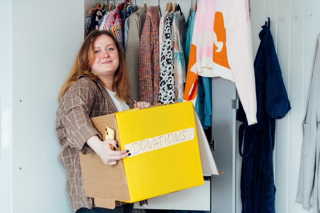 A woman smiles as she holds a box labeled “donations” in her closet.