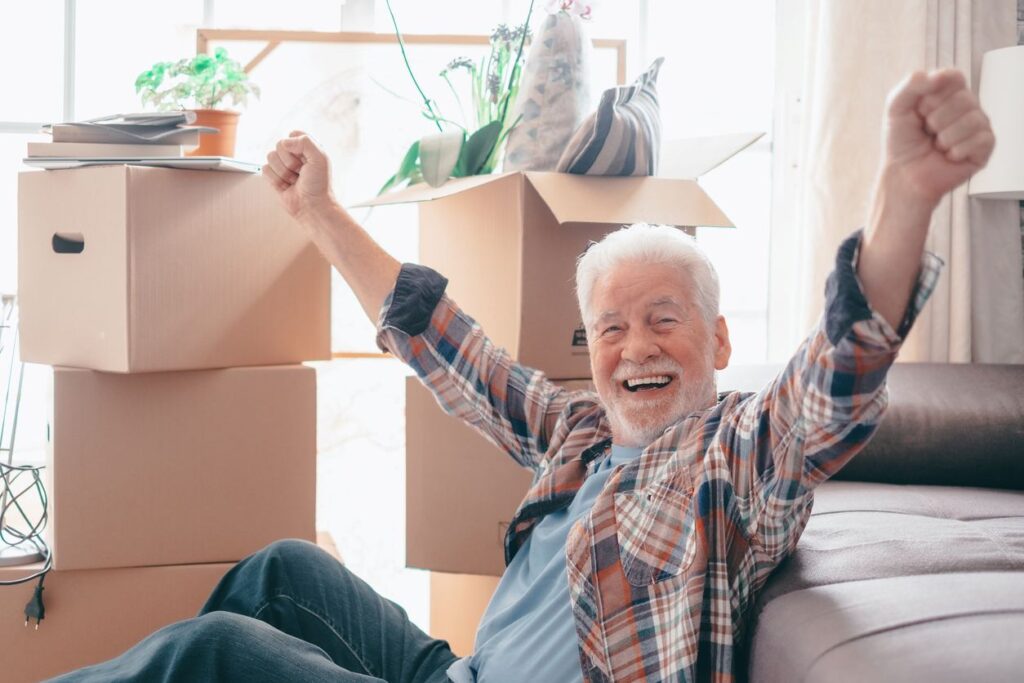 A senior smiles and celebrates with his arms in the air as he packs cardboard boxes before moving.