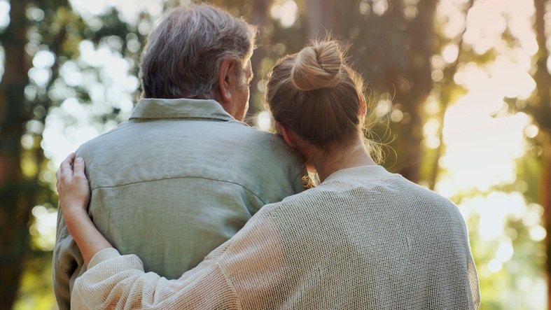 A father and his daughter are in the park, with her arm around his shoulder.