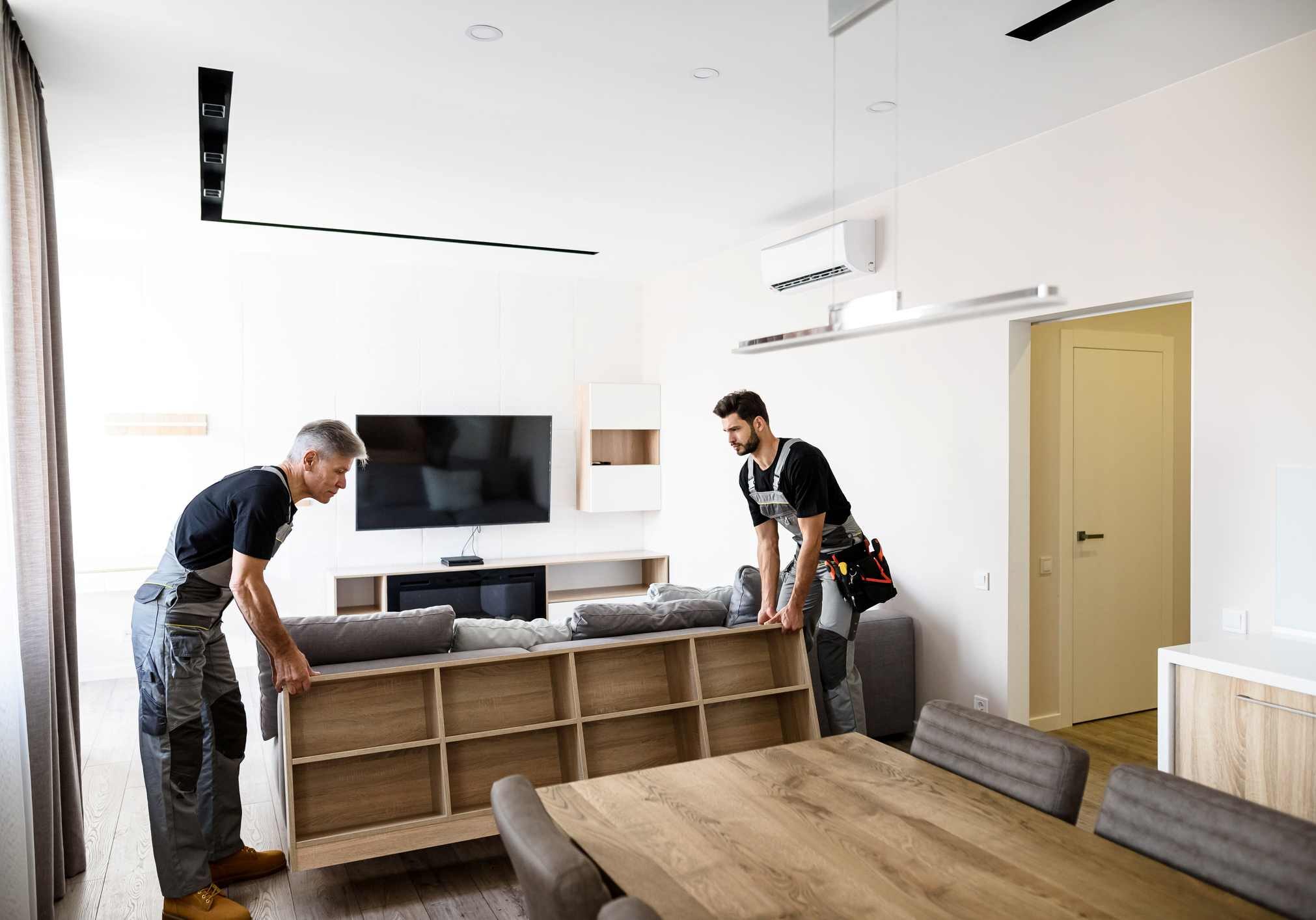 Full length shot of two contractor employees, young and aged worker in uniform moving personal belongings, furniture inside apartment. Moving service concept. Selective focus. Horizontal shot