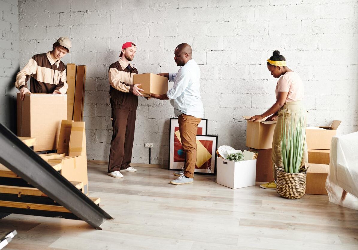 Two young workers in uniform helping married African couple with delivering packed boxes to new flat or house during remove