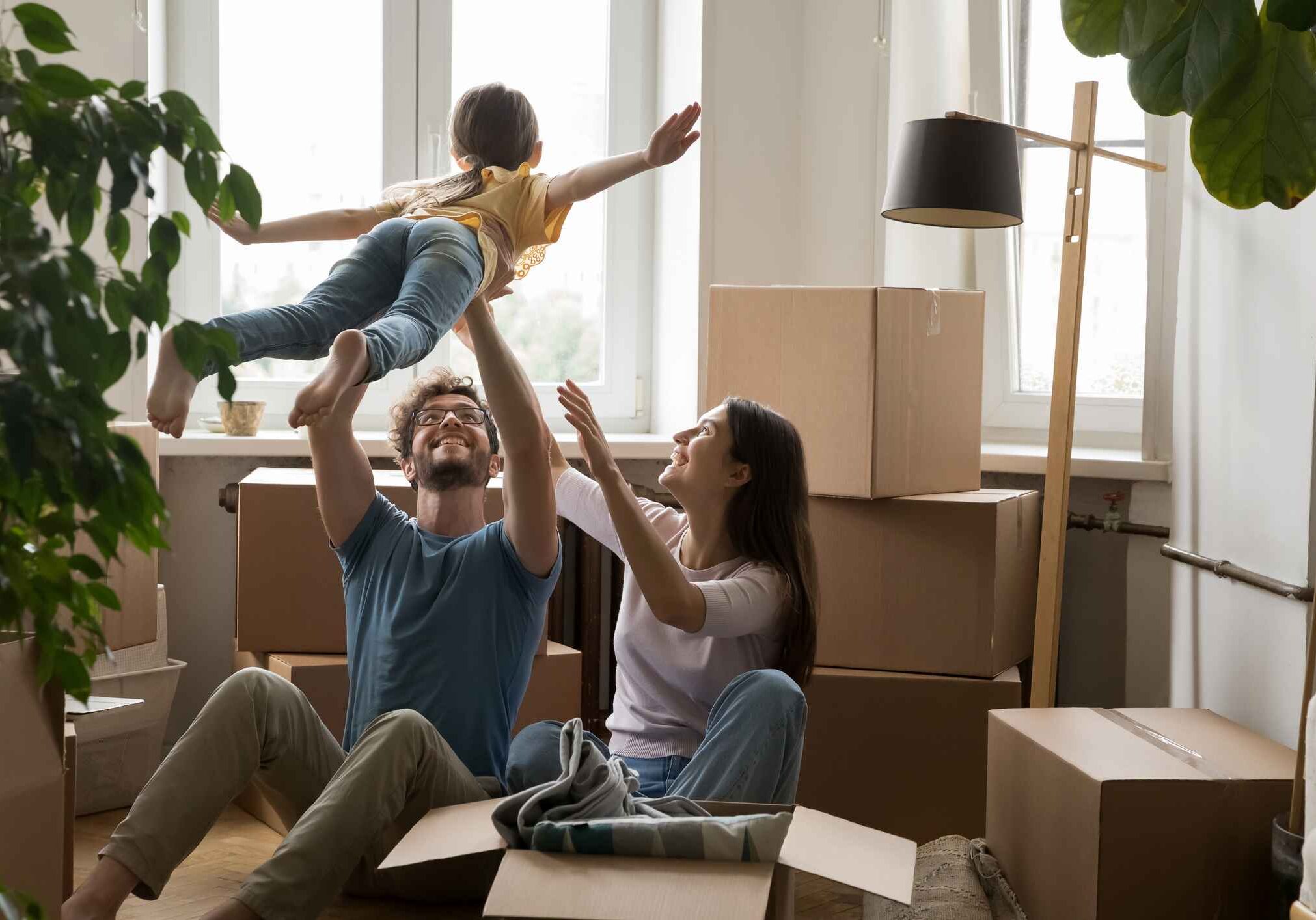 Cheerful young loving family with cute little daughter celebrate moving day, having fun, playing with cute little kid, feel happy at new house, sit on floor near heap of boxes. Bank loan, relocation