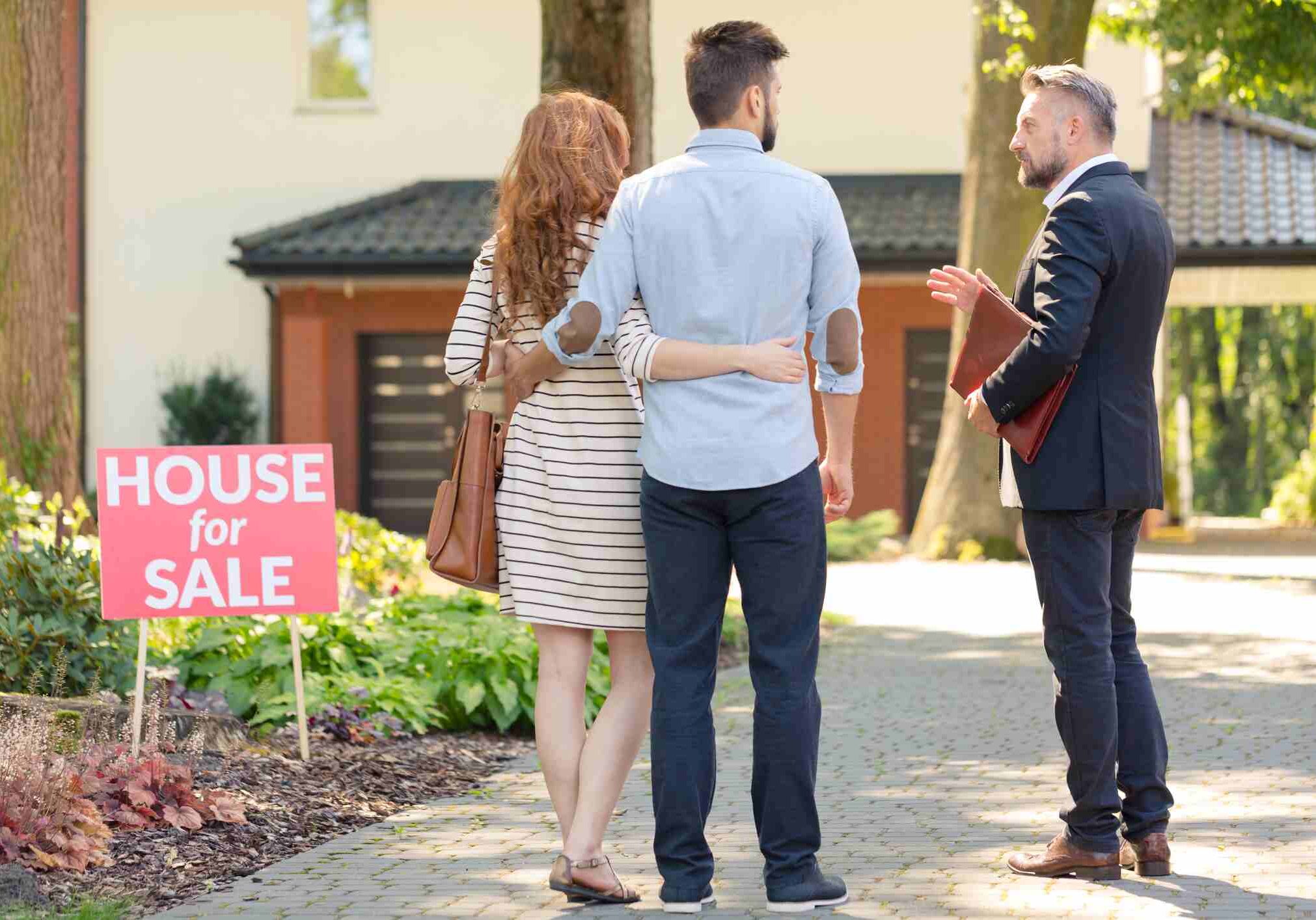 Real estate broker and clients standing next to plate with inscription "house for sale"