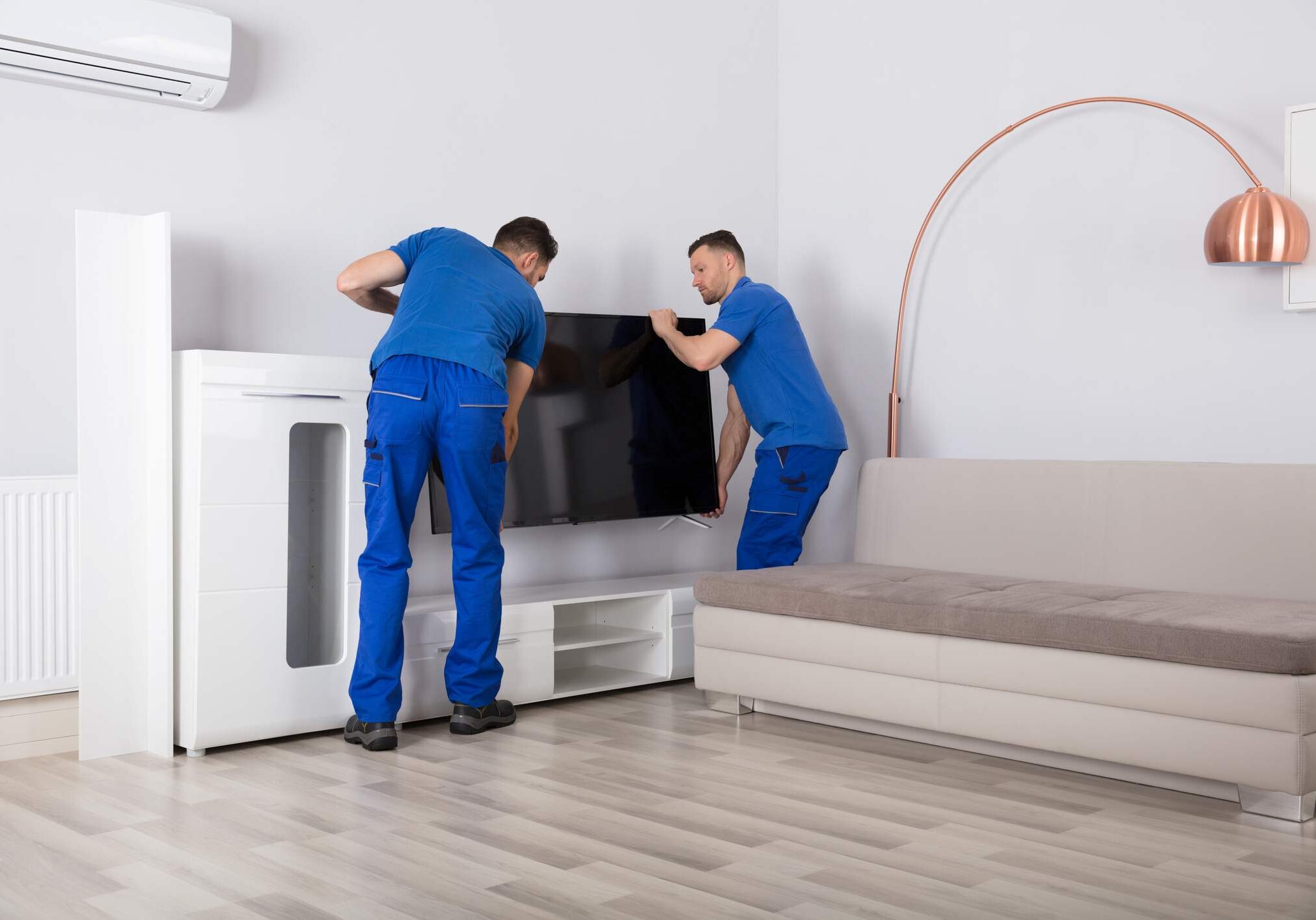 Two Young Male Movers In Uniform Placing Television In Living Room